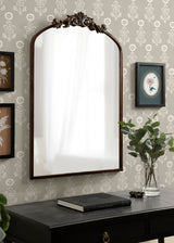 Wall-mounted arched mirror with dark wooden frame and ornate carved floral crest at top, reflecting a window, above a black table with book and glass vase with greenery.