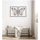 Two-piece framed art set depicting symmetrical neutral-toned watercolor butterfly wings on a subtle linen-textured background, hung above a wooden bench with draped fabric and open book.