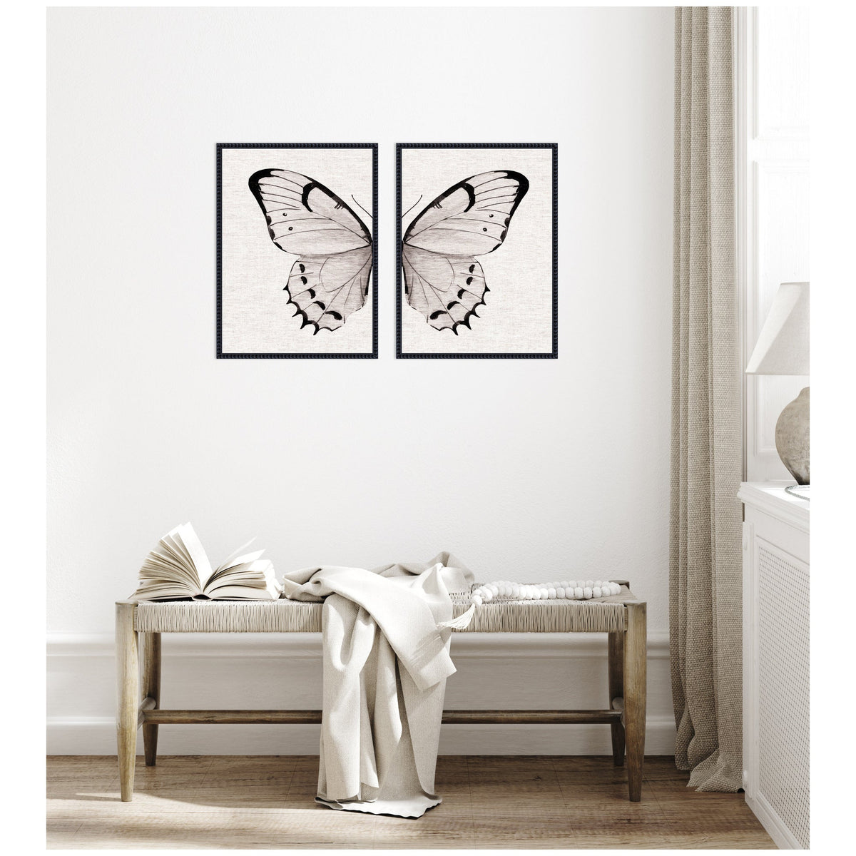 Two-piece framed art set depicting symmetrical neutral-toned watercolor butterfly wings on a subtle linen-textured background, hung above a wooden bench with draped fabric and open book.