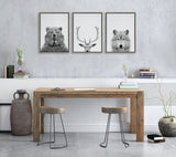 Three minimalistic framed animal portraits showing a bear facing front, a deer's head with antlers, and a wolf facing forward, hung on a gray textured wall above a wooden table with two stools.