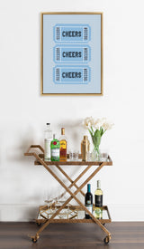 Wall art with three stacked blue tickets labeled 'CHEERS' above a wooden and glass bar cart holding liquor bottles, glasses, a shaker, and white tulips in a clear vase.