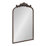 Tall rectangular mirror with an arched top and ornate dark brown carved floral crest framing the upper edge, reflecting a plain surface.