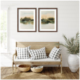 Two abstract landscape prints with muted greens and browns in dark wood frames above a wooden bench with three black-and-white gingham pillows, woven baskets, and a ceramic vase with greenery.