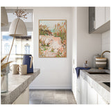 Framed pastel-toned painting depicting a sunrise seascape with beige rocks and greenish plants, hung on a white wall in a modern kitchen with gray countertops and white cabinets.