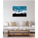 Framed aerial photo of white ocean waves breaking onto a black sand beach, hanging above a neutral-toned sofa with beige and striped pillows in a minimalist living room.