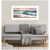 Horizontal painting of ocean waves with teal and white foam under a pastel pink and purple sky, framed in light wood, hanging above a gray sofa with gray pillows and throw.
