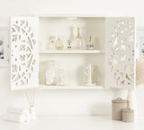 White carved wood wall cabinet with open doors revealing perfume bottles on top shelf and cotton balls with a cactus-shaped container on the bottom shelf, against a white wall.