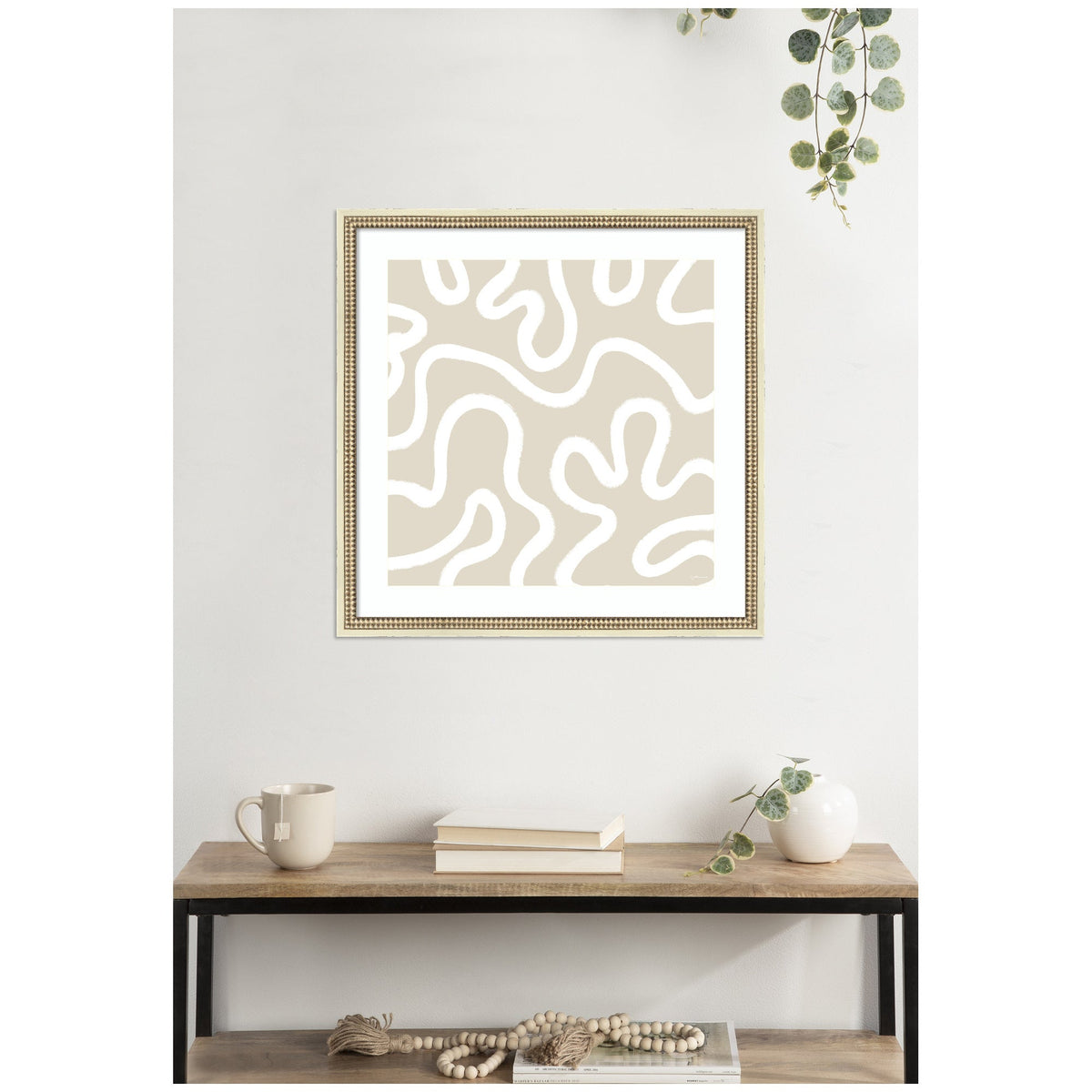Framed abstract wall art featuring beige background with white organic, wavy lines, displayed above a wooden shelf holding books, a mug, and a small white vase with greenery.