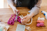Hands cutting pink yarn next to twisted white rope pieces and other colorful yarn bundles on a wooden table, with crafting tools and instructions nearby.