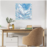Square framed canvas painting of a blue sky with fluffy white clouds, displayed above a wooden desk with beige upholstered chair and minimal decor.