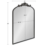 Tall rectangular mirror with a black metal frame topped by an ornate floral scrollwork arch, measuring 36 inches high and 24 inches wide.