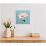 Square canvas with a pink daisy featuring a yellow center against a teal background, framed with a white scalloped edge, hanging above a wooden surface with plants and books.