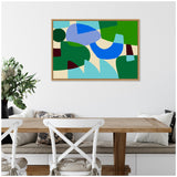 Abstract wall art with overlapping blue, green, teal, and beige shapes in organic and geometric forms, framed in light wood, hanging above a wooden dining table.