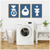 Three framed abstract ceramics-themed prints with white shapes on blue backgrounds hang above a modern white front-loading washing machine in a bright laundry room.