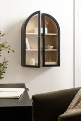 Black metal-framed wall cabinet with arched glass doors, light wood interior with three shelves holding white and neutral decorative items, mounted on a white wall.