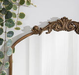 Upper corner of an Arendahl Traditional Arch Mirror with ornate scrollwork in antique bronze finish, next to cascading round green leaves of a plant on a white wall.