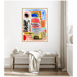 Framed abstract canvas with colorful shapes in orange, blue, black, pink, and green hangs above a wooden bench with an open book and a draped white cloth in a bright, minimalist room.