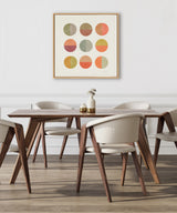 Square framed canvas featuring nine circles in warm earth tones and soft greens arranged in a 3x3 grid, hanging above a modern wood dining table with cream cushioned chairs.