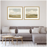 Two framed landscape prints depicting serene, cloudy skies over rolling beige hills, hung side by side above a gray sectional sofa with striped pillows in a minimalist living room.