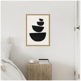 Framed canvas with black geometric shapes stacked vertically on a white wall above a wooden side table holding a white coffee cup and two books.