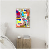 Abstract painting with vibrant blocks of red, blue, green, yellow, and brown, featuring bold black linear and spiral shapes in a natural wood frame on a white wall above a wood nightstand with books and white cup.