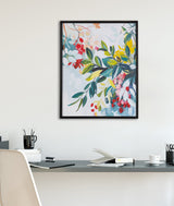 Framed botanical print with vibrant jewel-tone leaves and flowers in red, yellow, green, and pink on a light background hanging above a minimalist desk.
