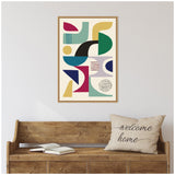 Framed abstract canvas with colorful geometric shapes in teal, mustard, black, and burgundy, hanging above a rustic wooden bench with an open book and a beige pillow saying 'welcome home'.