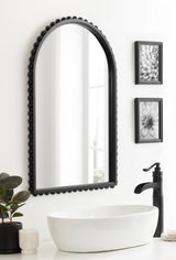 Arched wall mirror with black beaded frame hanging above a white oval vessel sink and black faucet, flanked by two black-framed flower photos on a white wall.