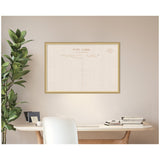 Framed fabric pinboard with vintage linen postcard print hanging above a wooden desk with stacked books and a green leafy plant to the left.