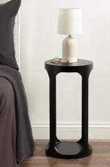 Dark round wooden drink table with an open rectangular center, holding a white ceramic lamp with beige shade and a pair of eyeglasses, beside a gray fabric sofa.