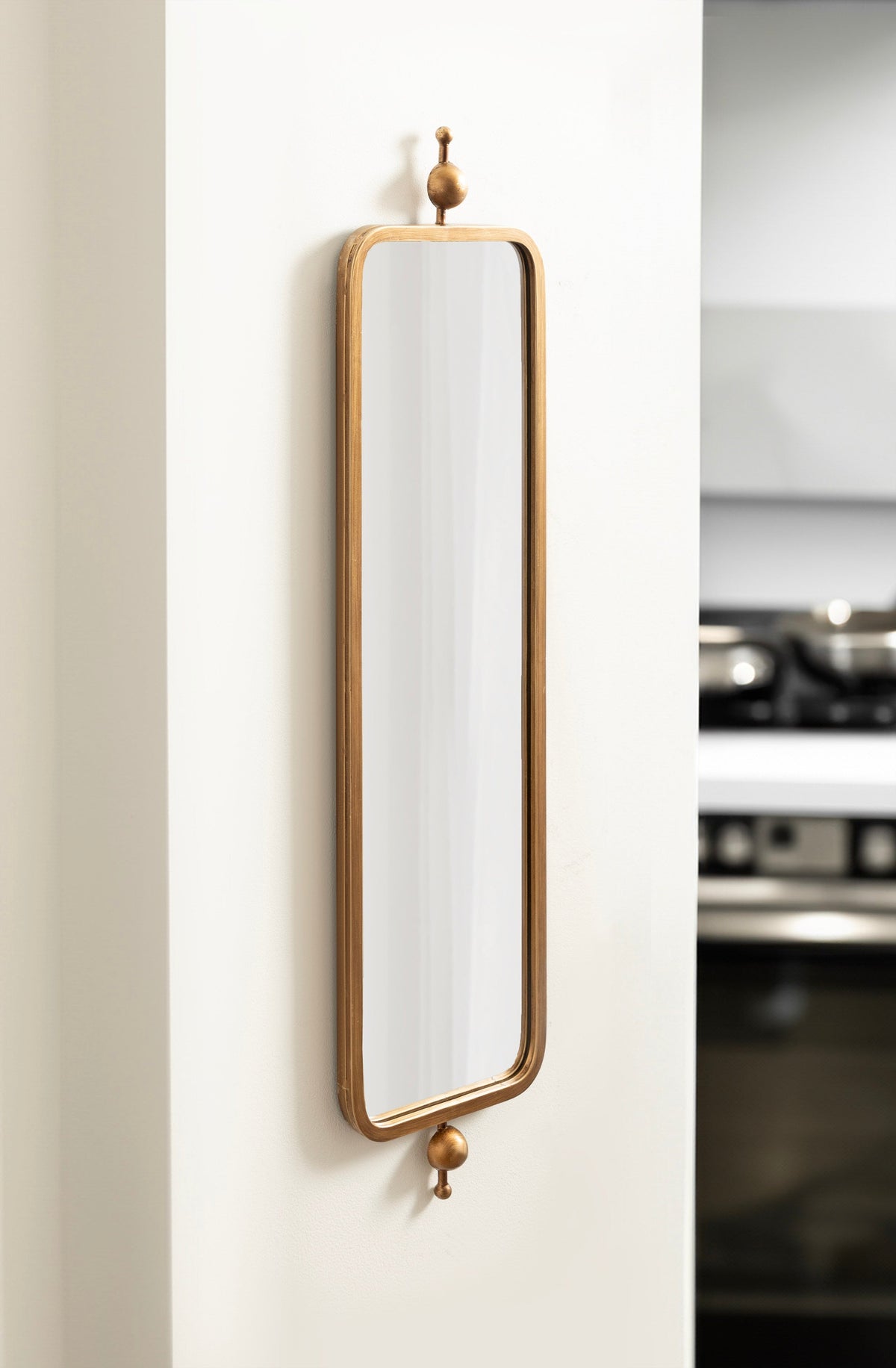 Tall, narrow rectangular mirror with rounded corners and a warm wooden frame, mounted vertically on a white wall with decorative wooden finials top and bottom.