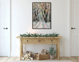 Framed canvas painting of a sunlit forest path with tall trees in soft pastel tones of pink, green, and beige, hanging above a wooden console table with pine garland and candles.