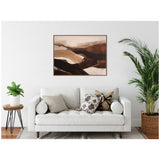 Abstract landscape art with layered brown, beige, and black tones in a wood frame above a white sofa with patterned cushions flanked by green plants in a modern living room.
