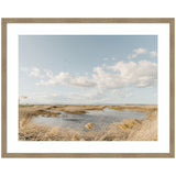 Framed photograph of a marshland with golden grasses surrounding a winding water channel under a partly cloudy blue sky with a small flock of birds flying above.