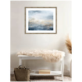 Framed abstract art with tonal blues and soft beige, resembling an oceanic scene, hanging above a white bench with a fluffy beige throw and a green bag on the floor.