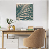 Square canvas print of teal and beige maximalist palm leaves with a modern retro style, framed in light wood, hanging above a wooden desk and tan upholstered chair.