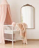 White baby crib draped with a pink canopy and a peach blanket hanging on the side, next to a tall decorative wall mirror with a carved bronze top frame.