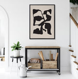 Abstract black distorted shapes on tan background in a black frame hanging above a rustic wood and metal console table with wooden crates and a bag underneath, beside a small plant on a round black and white side table.