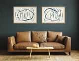 Two framed abstract canvas artworks with expressive blue and green looping lines, hung side by side on a dark teal wall above a brown leather sofa.
