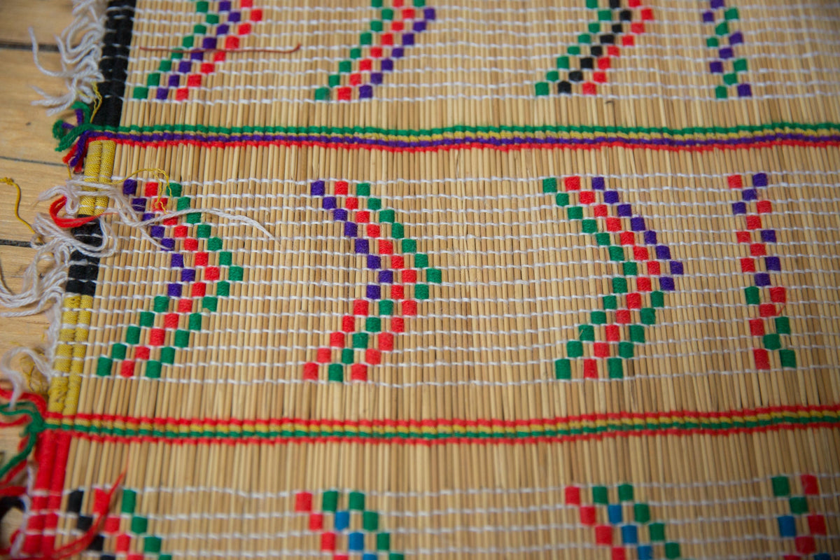 Close-up of a woven mat with diagonal arrow patterns in green, red, and purple on a natural beige background with colorful fringed edges.