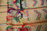 Close-up of a woven mat with colorful geometric patterns in red, green, purple, and yellow, featuring fringed edges and a light natural straw base on wooden floorboards.