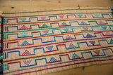 Woven vintage African Tuareg rug mat with colorful geometric triangle and step patterns in red, blue, green, and purple on natural beige fibers, laid flat on wood surface.