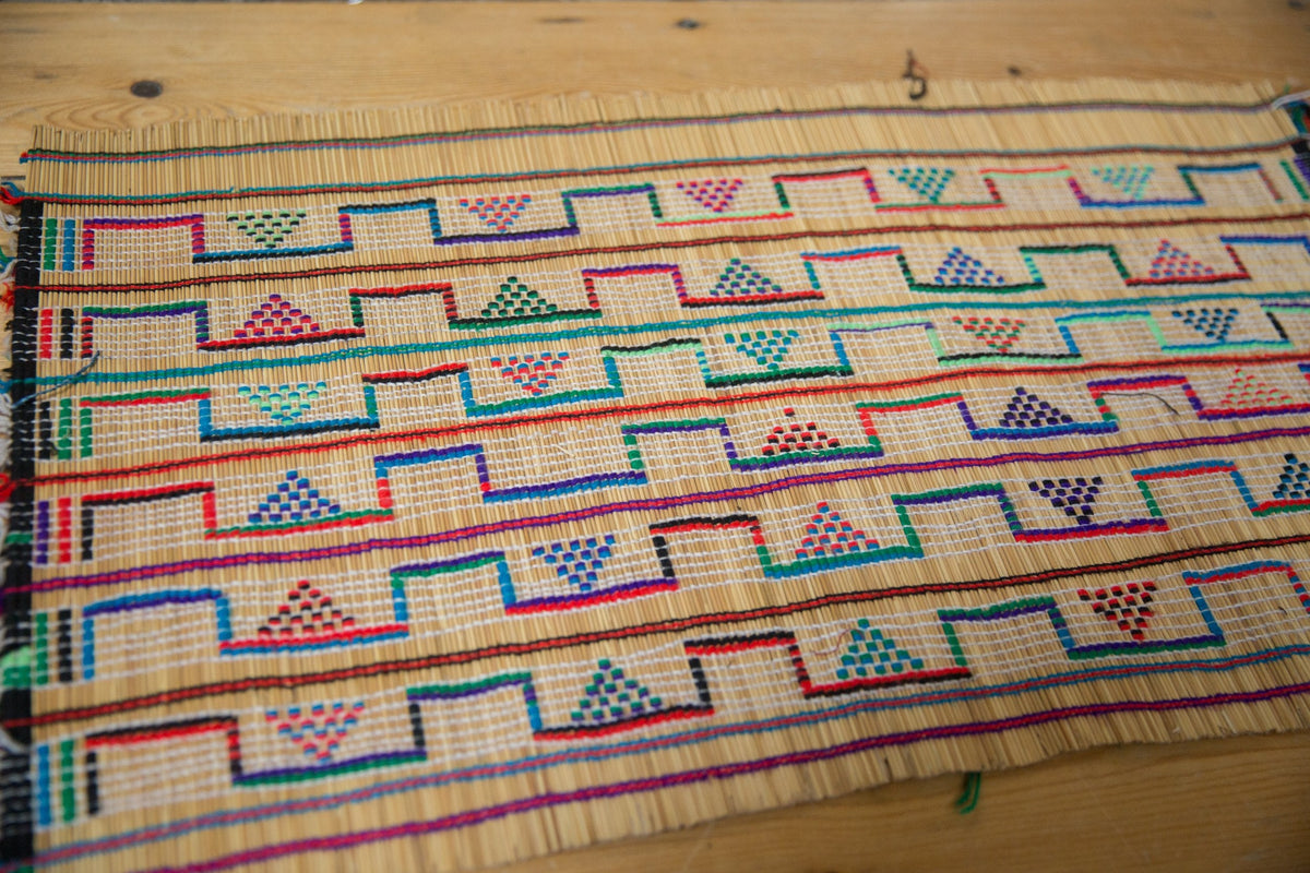 Woven vintage African Tuareg rug mat with colorful geometric triangle and step patterns in red, blue, green, and purple on natural beige fibers, laid flat on wood surface.