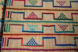 Close-up of a woven mat featuring repeated geometric patterns with colorful triangular and rectangular shapes in red, green, blue, and black on a natural beige background.