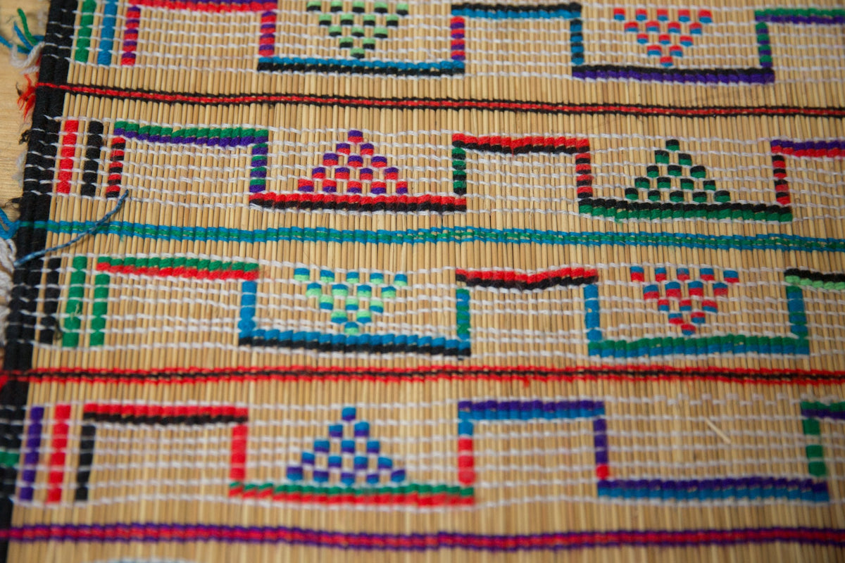 Close-up of a woven mat featuring repeated geometric patterns with colorful triangular and rectangular shapes in red, green, blue, and black on a natural beige background.