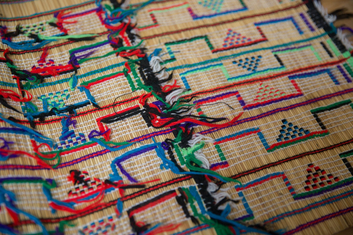 Close-up of a woven rug with a grid pattern containing small triangle designs in red, blue, and green, accented by colorful tassels and black outlines.