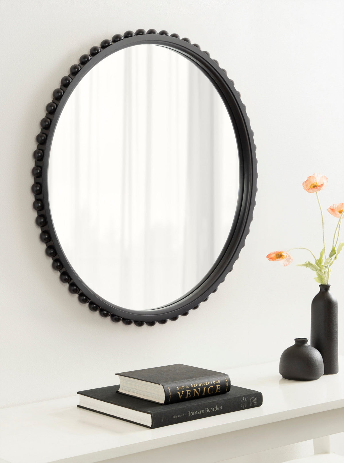 Round wall mirror with a black frame featuring evenly spaced bead-like accents, hanging above a white surface with black vases and stacked books.