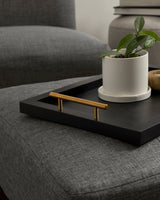 Black rectangular tray with gold handles placed on gray fabric furniture, holding a small white ceramic plant pot with green leaves.