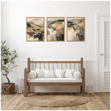 Three framed abstract landscape paintings with earthy tones above a wooden bench with white cushions in a minimalist living room featuring a woven rug and potted plant.