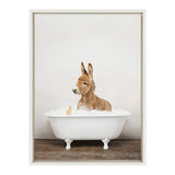 Young brown donkey sitting in a white clawfoot bathtub filled with bubbles, accompanied by a small yellow rubber duck on a wooden floor with a plain light wall behind.